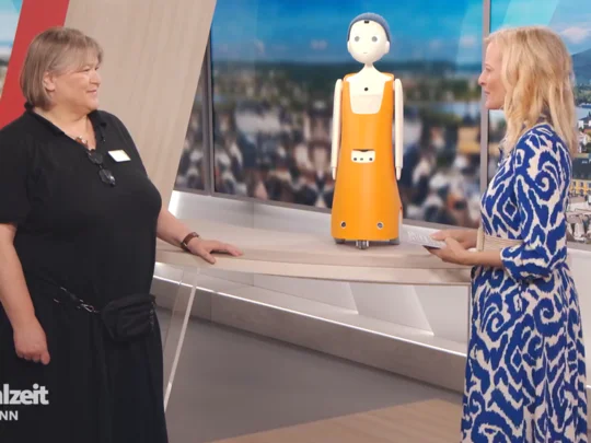 Betreuungskraft mit Sozialroboter wird im Fernsehstudio interviewt